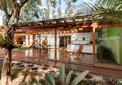 Fotografia de ambiente externo de casa tropical, com piso e móveis de madeira e diversas plantas.